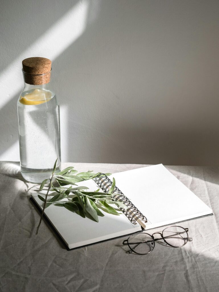 Simple composition with open notebook, eyeglasses, and water bottle on sunlit table.